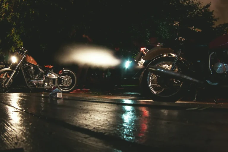 Cruiser motorcycles parked on wet pavement at night, a headlight streaking past