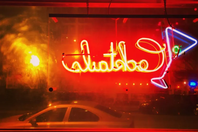 Red neon cocktail sign glowing in a bar window at night, a car passing on the street outside