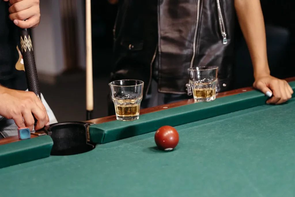 Whiskey glasses on the rail of a pool table while players in leather chalk their cues