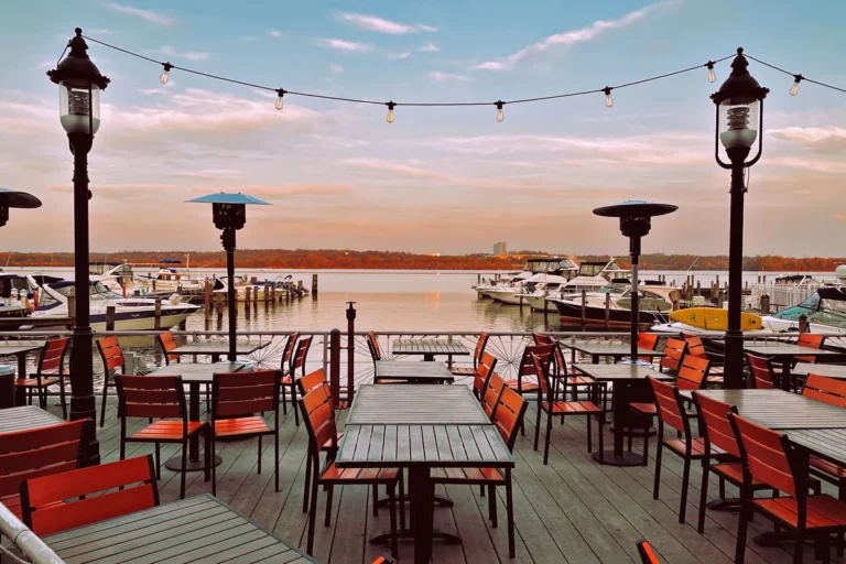 Waterfront deck with tables, string lights and patio heaters overlooking a marina at sunset