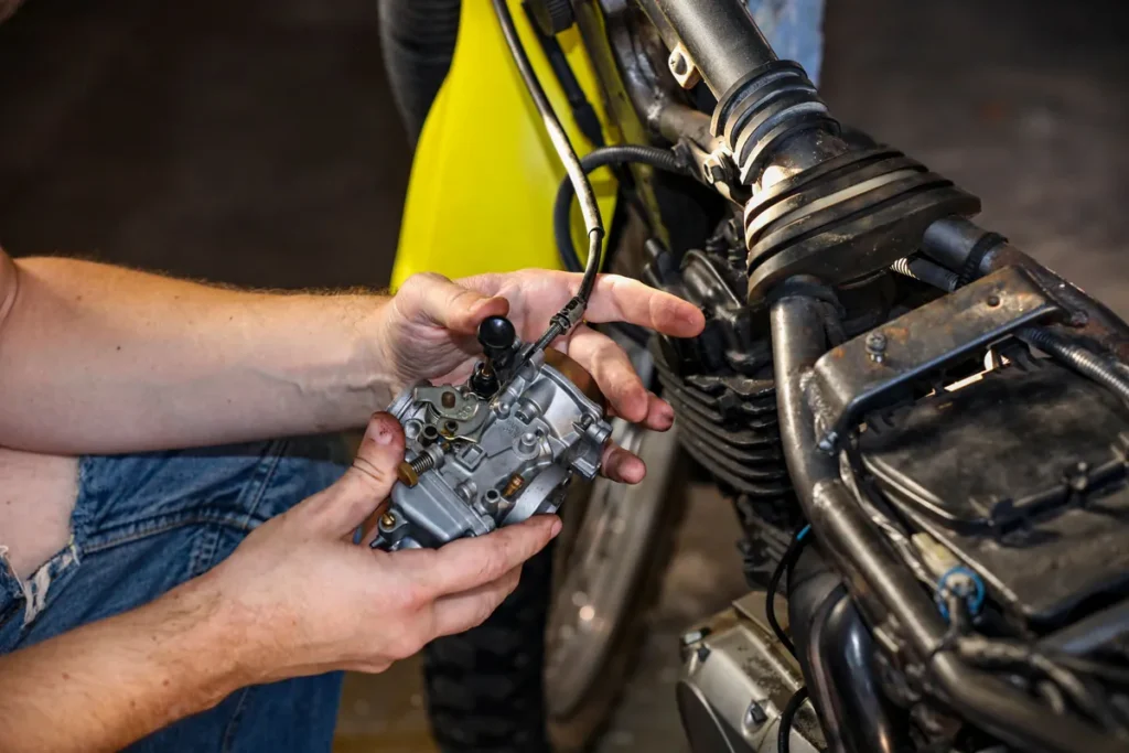 Mechanic's hands holding a carburetor beside a motorcycle under repair