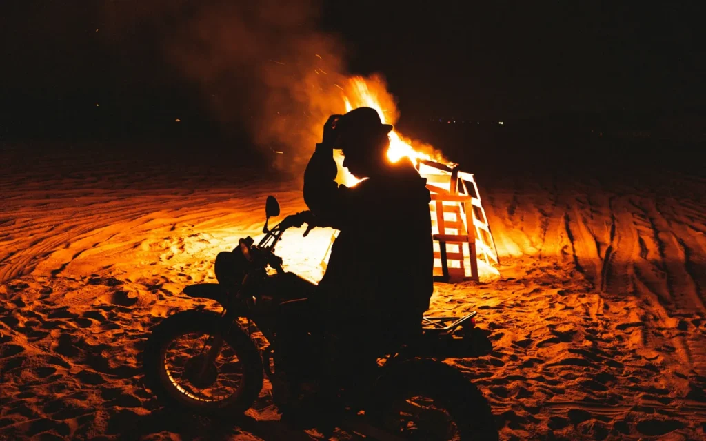Rider silhouetted on a motorcycle beside a blazing bonfire on a beach at night