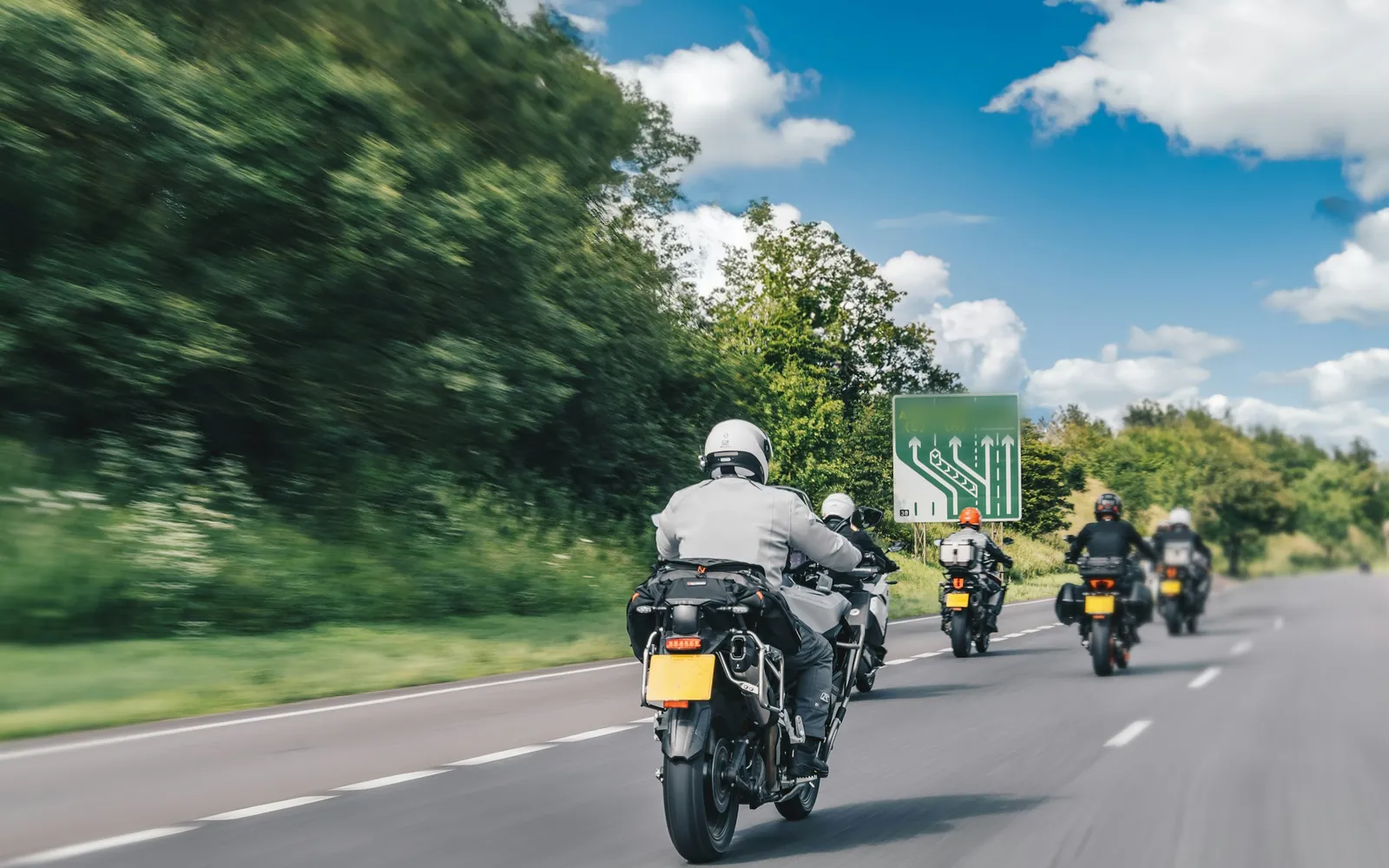Five touring motorcycles riding in a line down an open two-lane road on a summer day, seen from behind