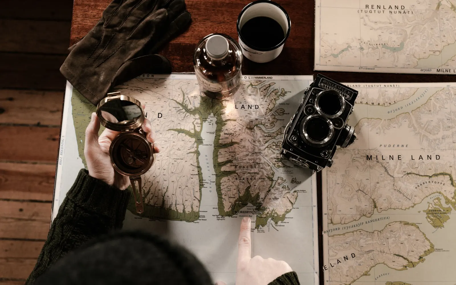 Trip planning on a wooden table: maps, an enamel mug of coffee, leather gloves, a compass and a camera