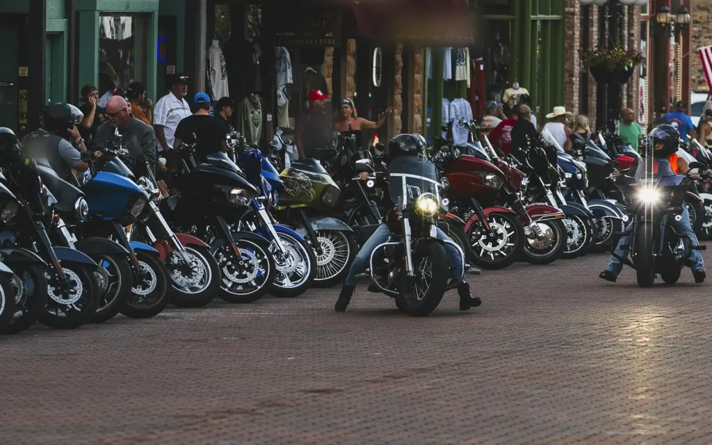 Rows of touring motorcycles parked along a brick main street as riders roll past at a summer rally