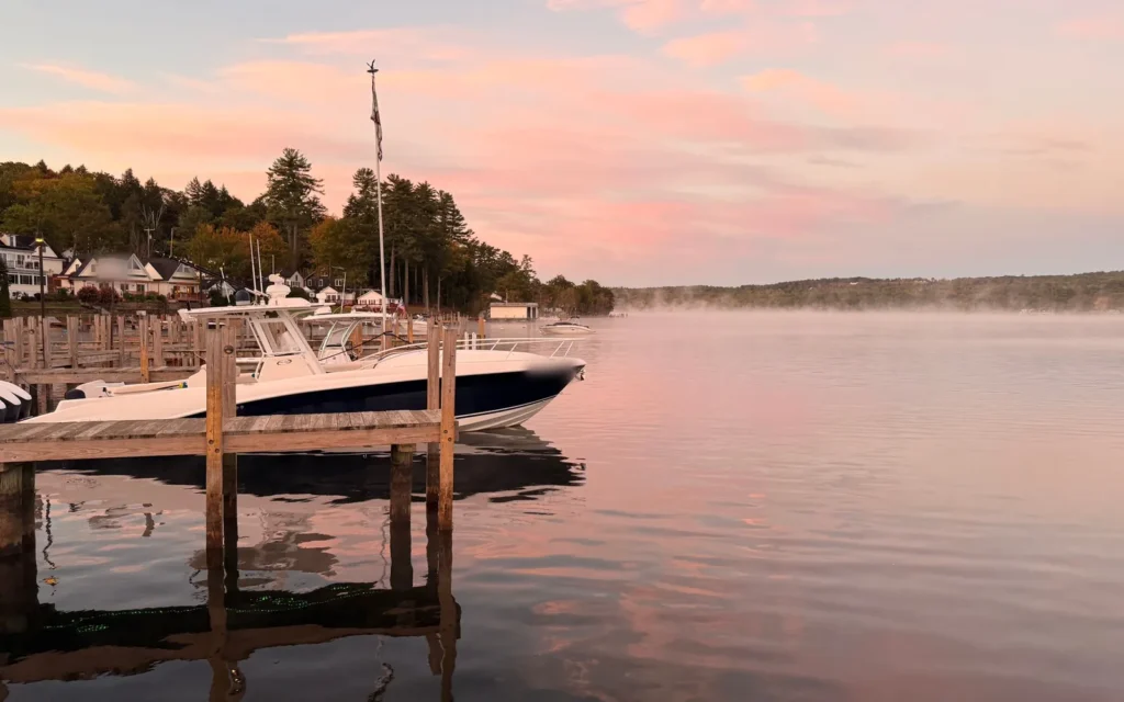 Boats moored at a wooden dock under a pink sunset sky, mist drifting over Lake Winnipesaukee, NH