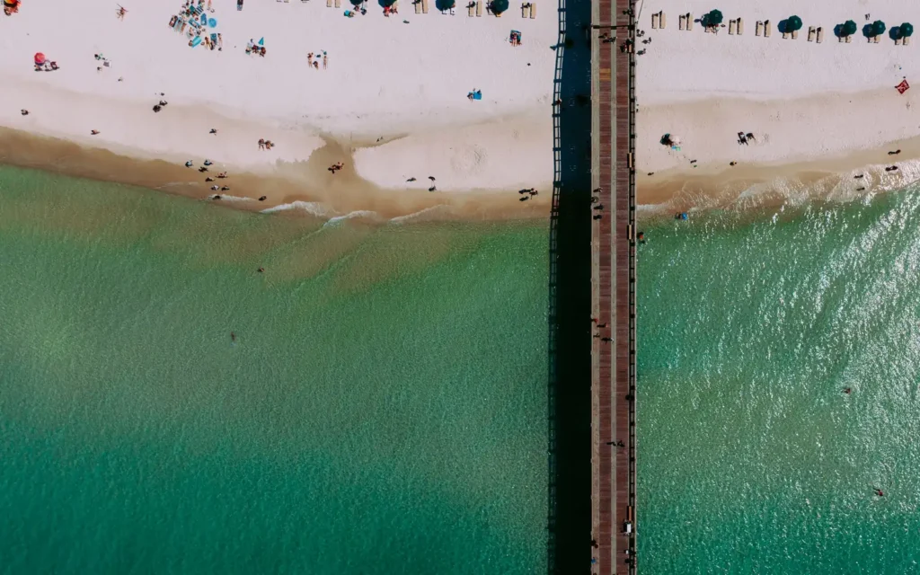 Top-down aerial of a pier crossing white sand into emerald Gulf water at Panama City Beach, FL