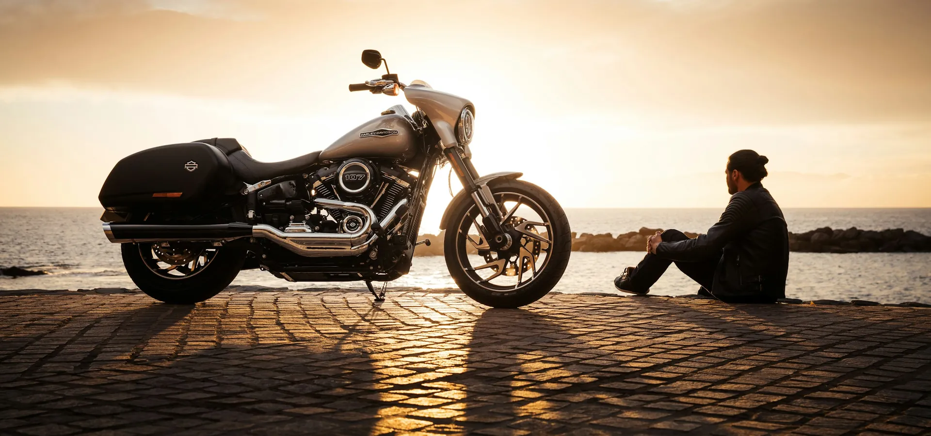 Cruiser motorcycle silhouetted by the sea at golden hour, a rider resting beside it, the cover photo of our Biketoberfest guide