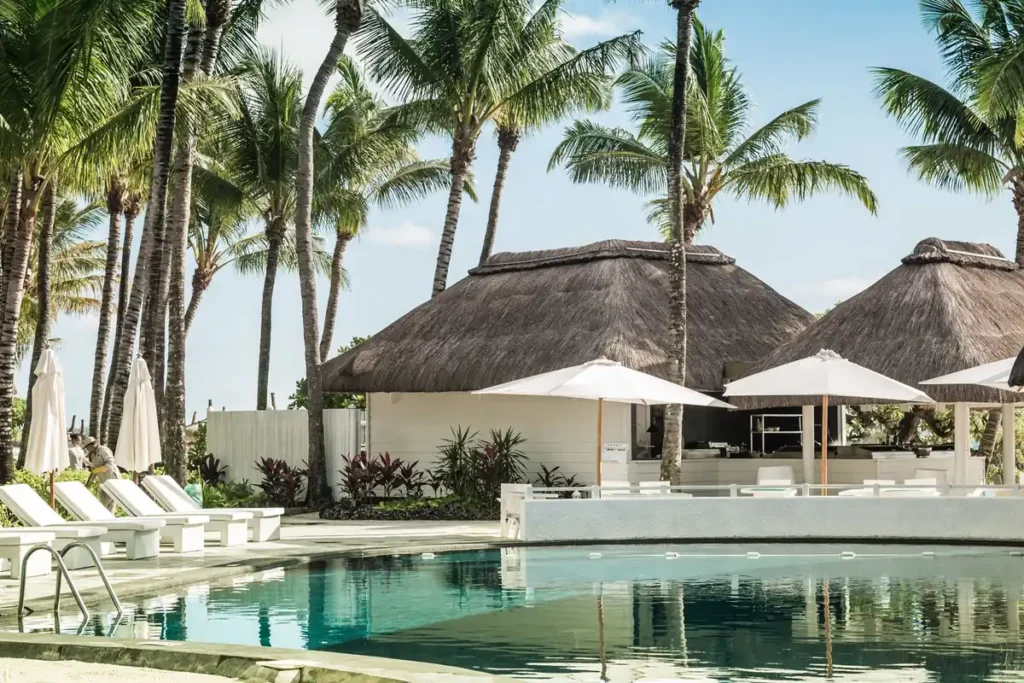 Resort pool with sun loungers, umbrellas and thatched cabanas under tall palm trees